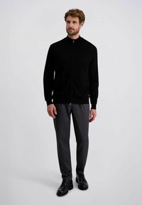 Schwarzer Reißverschluss-Pullover mit hohem Kragen, kombiniert mit strukturierten, gemusterten grauen Hosen. Das Modell trägt schwarze Schuhe. Schlichter Hintergrund.