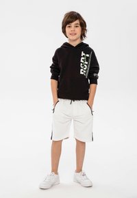 Sudadera negra con capucha y estampado gráfico, combinada con pantalones cortos blancos que tienen franjas laterales negras y bolsillos. Zapatillas blancas completan el look.