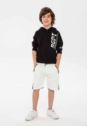 Zwarte sweatshirt met capuchon en grafische print, gecombineerd met witte shorts met zwarte zijstrepen en zakken. Witte sneakers maken de look compleet.