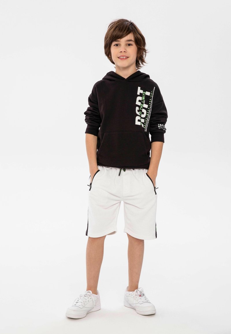 Sudadera negra con capucha y estampado gráfico, combinada con pantalones cortos blancos que tienen franjas laterales negras y bolsillos. Zapatillas blancas completan el look.