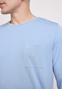 Mand iført en lyseblå langærmet skjorte med en brystlomme, synlig fra halsen til midten af torsoen, mod en ensfarvet baggrund.