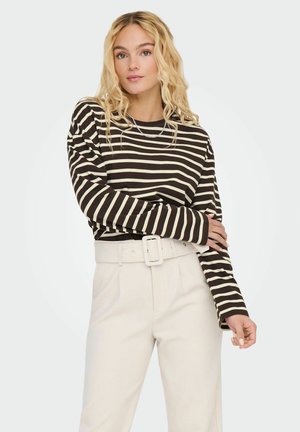 Jeune femme aux longs cheveux blonds portant un t-shirt à manches longues rayé noir et blanc et un pantalon taille haute beige avec une ceinture large.