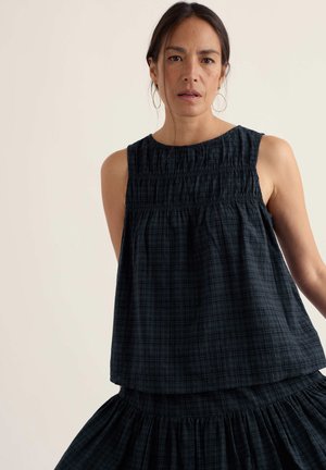 Kobieta w ciemnym, bezrękawnym topie w kratę i pasującej spódnicy, z dużymi kolczykami kołami, stoi na tle jednolitego jasnego tła.