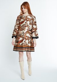 Braunes und beiges gemustertes Kleid mit langen Ärmeln, das einen runden Ausschnitt und einen dekorativen Saum hat. Dazu passende beigefarbene Stiefel bis zum Knie.