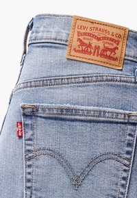 Jean en denim bleu clair avec une finition texturée, comportant une pièce en cuir marron et une étiquette rouge sur la poche arrière, présentant des motifs de couture classiques.