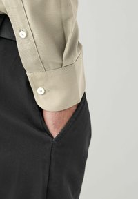 Beige pamut ing ujja gomb részletekkel a mandzsettán, fekete szövetnadrággal párosítva, oldalzsebbel. A textúrázott anyag jól látható.