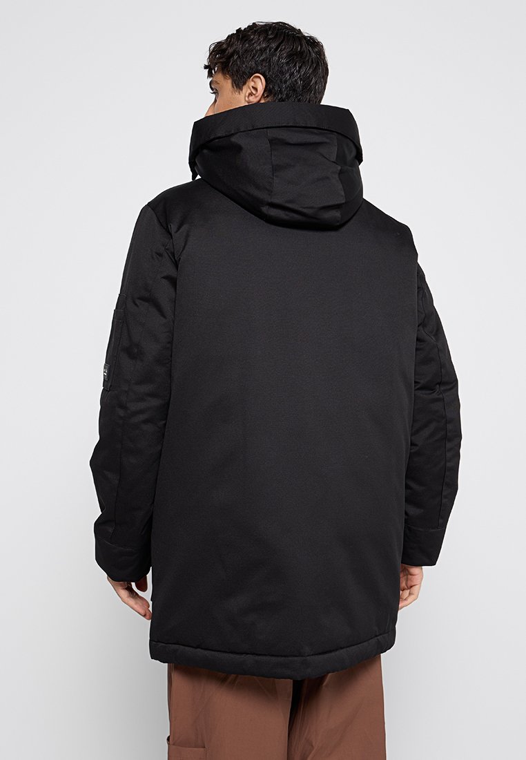 Parka noire avec une capuche spacieuse. Présente une texture lisse et mate, des manches longues et un ourlet droit. Design minimaliste sans ornements.