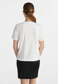 Biała bawełniana koszulka z krótkim rękawem i okrągłym dekoltem, zestawiona z czarną dopasowaną spódnicą, widok z tyłu. Gładka faktura materiału.