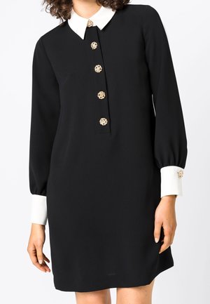 Musta pitkähihaisen mekko, jossa valkoinen kaulus ja kalvosimet, koristeltu viidellä koristeellisella kultanapilla edessä ja kalvosimissa.