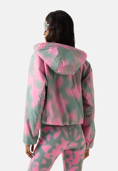 Kvinde iført lyserød og grøn camouflage hættetrøje og matchende leggings, set bagfra med langt mørkt hår og solbriller.