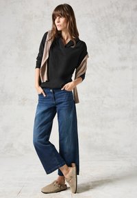 Schwarz v-ausschnitt Langarmshirt, beiger drapierter Pullover, blaue weit geschnittene Jeans und beige Clogs vor einem strukturierten neutralen Hintergrund.