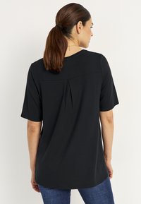 Svart skjorta med korta ärmar, rund halsringning och detalj med veck i ryggen. Kombinerad med blå jeans. Slät tygtextur.