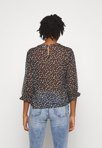 Blusa preta transparente com pequenos padrões florais, mangas três quartos e abertura em forma de chave nas costas. Tecido leve com um design fluido.