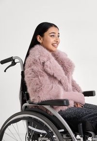 Cappotto rosa in pelliccia sintetica con un ampio colletto, abbinato a un maglione rosa a coste. Modella seduta su una sedia a rotelle, indossa pantaloni neri con motivo.