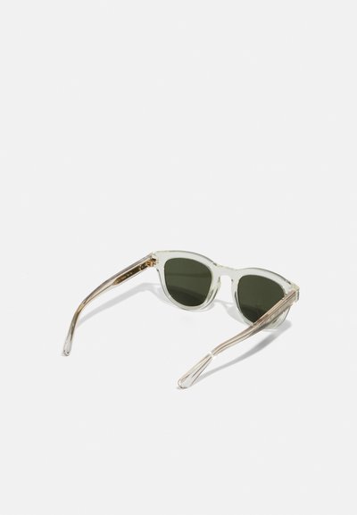 Óculos de sol com armação transparente e lentes verdes, em formato de olho de gato, e braços transparentes. Detalhes texturizados notáveis na área das hastes.