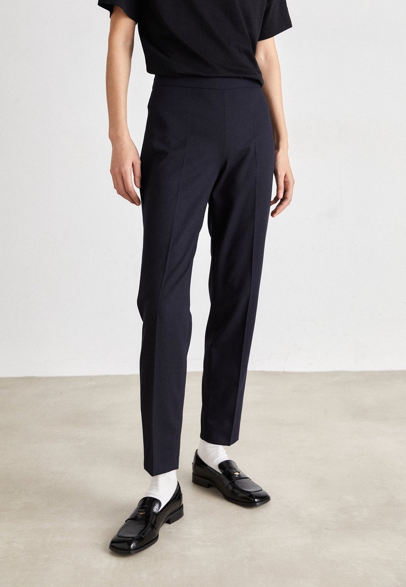 Navy tailored trousers with a slim fit, featuring a smooth texture, front creases, and a mid-rise waist. Paired with black loafers and white socks.