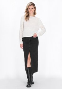 Femeie îmbrăcată cu un pulover alb din tricot, fustă neagră până la talie cu slit frontal și cizme negre până la genunchi, stând cu mâna pe șold.