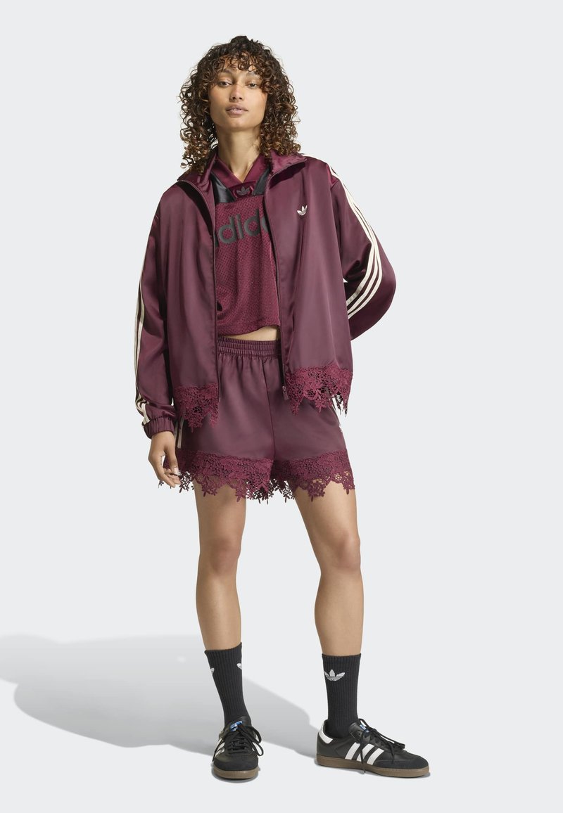Femme portant une veste Adidas bordeaux, un crop top en résille, un short bordé de dentelle, des chaussettes noires et des baskets Adidas noires, debout devant un fond uni.