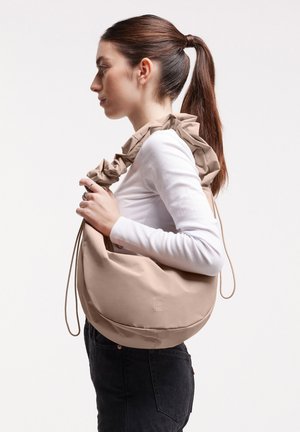 Donna con coda di cavallo marrone che indossa una maglietta bianca e pantaloni neri, con una grande borsa beige a tracolla con manico arricciato.