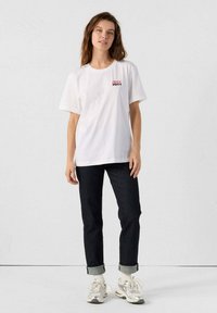 Camiseta de algodón blanca con mangas cortas, que presenta un gráfico en rojo y rosa "JOTT" en el pecho. Combinada con jeans de mezclilla oscura y zapatillas.