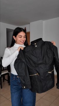 Γυναίκα με λευκό ψηλό γιακά και τζιν κρατάει ένα μαύρο σακάκι με χρυσά φερμουάρ και κουμπιά, χαμογελώντας μέσα σε ένα δωμάτιο με καρέκλες.