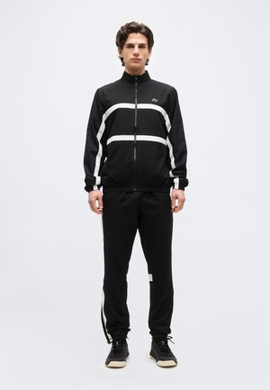 Mladý muž stojaci, oblečený v čierno-bielej športovej bunde na zips a zodpovedajúcich nohaviciach s čiernymi teniskami na jednoliatom pozadí.