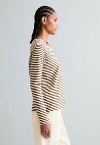 Frau mit geflochtenem Haar, die ein braun-weiß gestreiftes Langarmshirt und cremefarbene Hosen trägt und im Profil vor einem schlichten hellen Hintergrund steht.