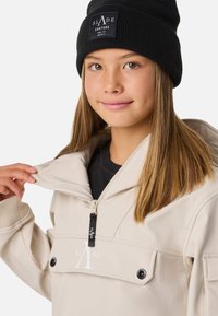 Giacca con cappuccio color crema con tasca con zip, cursore della zip nero e dettaglio con logo. Indossata sopra una maglia nera, abbinata a un berretto nero.