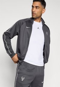 Gray athletic jacket with white piping, featuring Nike logo. Worn over a white shirt with a black Jordan logo, paired with gray pants.
