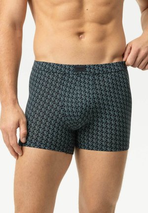 Männlicher Torso trägt dunkle Boxershorts mit hellem abstraktem Muster und passt den Bund auf der rechten Seite vor einfarbigem Hintergrund an.