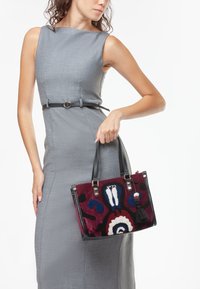 Borsa in bordeaux e nera, con motivi floreali blu, bianchi e rossi. Realizzata in tessuto testurizzato con accenti in pelle nera.