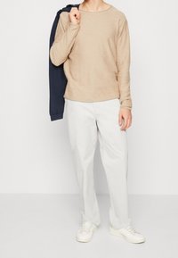 Beige långärmad skjorta med rund hals, i kombination med vita raka byxor och vita sneakers; en marinblå jacka hängande över ena axeln.