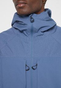 Niebieska, lekka kurtka z wysokim kołnierzem, teksturowanym korpusem, dwiema zapinanymi na zamek kieszeniami i sznurkami do regulacji dopasowania.