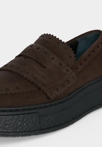 Mocassin en daim marron avec motifs décoratifs perforés et semelle noire épaisse texturée sur fond blanc.