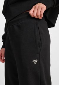 Schwarzer Sweatshirt mit ribbeltem Kragen, kombiniert mit schwarzen Jogginghosen, die seitliche Taschen und ein kleines weißes gesticktes Logo am Bein haben.