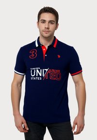 Polo din bumbac de culoare navy cu guler contrastant roșu, logo brodat și design de text alb. Are mâneci scurte și o croială ajustată.
