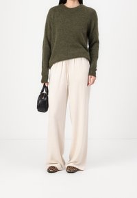 Maglione lavorato a maglia verde oliva, pantaloni beige a gamba larga, borsa nera e scarpe con stampa leopardata. Texture morbide e vestibilità rilassata.