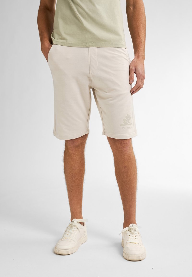 Homme portant un short décontracté beige avec poches et des baskets beige clair, se tenant avec une main dans la poche contre un fond uni.