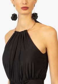 Schwarzes Neckholder-Kleid mit geraffter Textur am Ausschnitt. Das Model trägt große schwarze Blumenohrstecker und eine dünne goldene Kette.