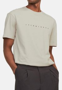 Lys beige bomulds t-shirt med rund halsudskæring, korte ærmer og "JACK&JONES" trykt i sort over brystet. Enkel design.