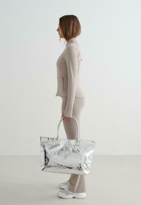 Metallisch silberne Tragetasche mit glänzender Oberfläche, doppelten Griffen und geprägtem Text. Das Model trägt ein beiges, figurbetontes Outfit und silberne Sneakers.