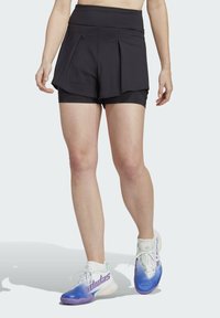 Sort træningsshorts med et lagdelt design, som har en høj talje og ydre plisseringer. Sammen med gradient blå og hvide sneakers.