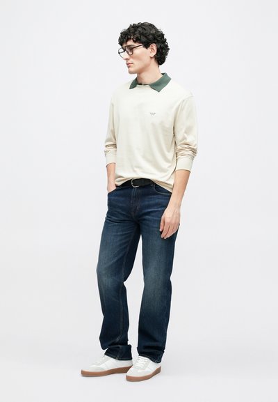 Gai krēmkrāsas garām piedurknēm krekls ar zaļu apkakli, kombinēts ar tumši zilām džinsu biksēm un baltām sporta kurpēm, krekla priekšā ar smalku logotipu.