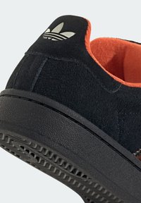 Černé semišové tenisky s oranžovým interiérem. Mají texturovanou černou gumovou podrážku a stříbrný logo akcent na patě.