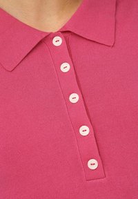 Primo piano di un colletto di una polo rosa lavorata a maglia con cinque bottoni bianchi allineati verticalmente sulla patta.