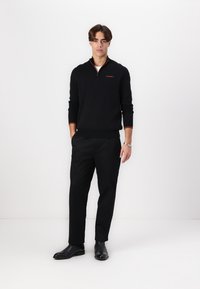 Suéter negro con cuello de cremallera y detalle de logo naranja, combinado con pantalones y zapatos negros. Textura suave, diseño ajustado y esquema de color monocromático.