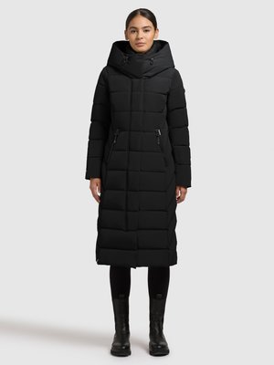 Zwarte lange gewatteerde jas met capuchon, voorzien van doorgestikte panelen, voorzakken met rits en een aansluitend silhouet. Draagt over zwarte leggings en laarzen.