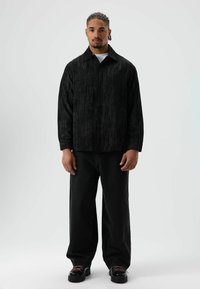 Zwart textuur shirt met een spitse kraag, lange mouwen en een iets losse pasvorm, gecombineerd met zwarte wijde broekspijpen en stevige zwarte schoenen.