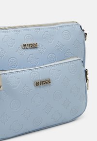 Deux pochettes zippées Guess bleu clair avec motifs en relief et plaques de logo dorées sur fond blanc.