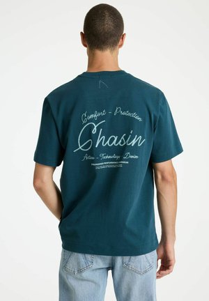 Man draagt een teal gekleurd T-shirt met korte mouwen met de tekst "Comfort - Protection Chasin Active - Technology - Denim", gecombineerd met lichtblauwe jeans, van achteren gezien.